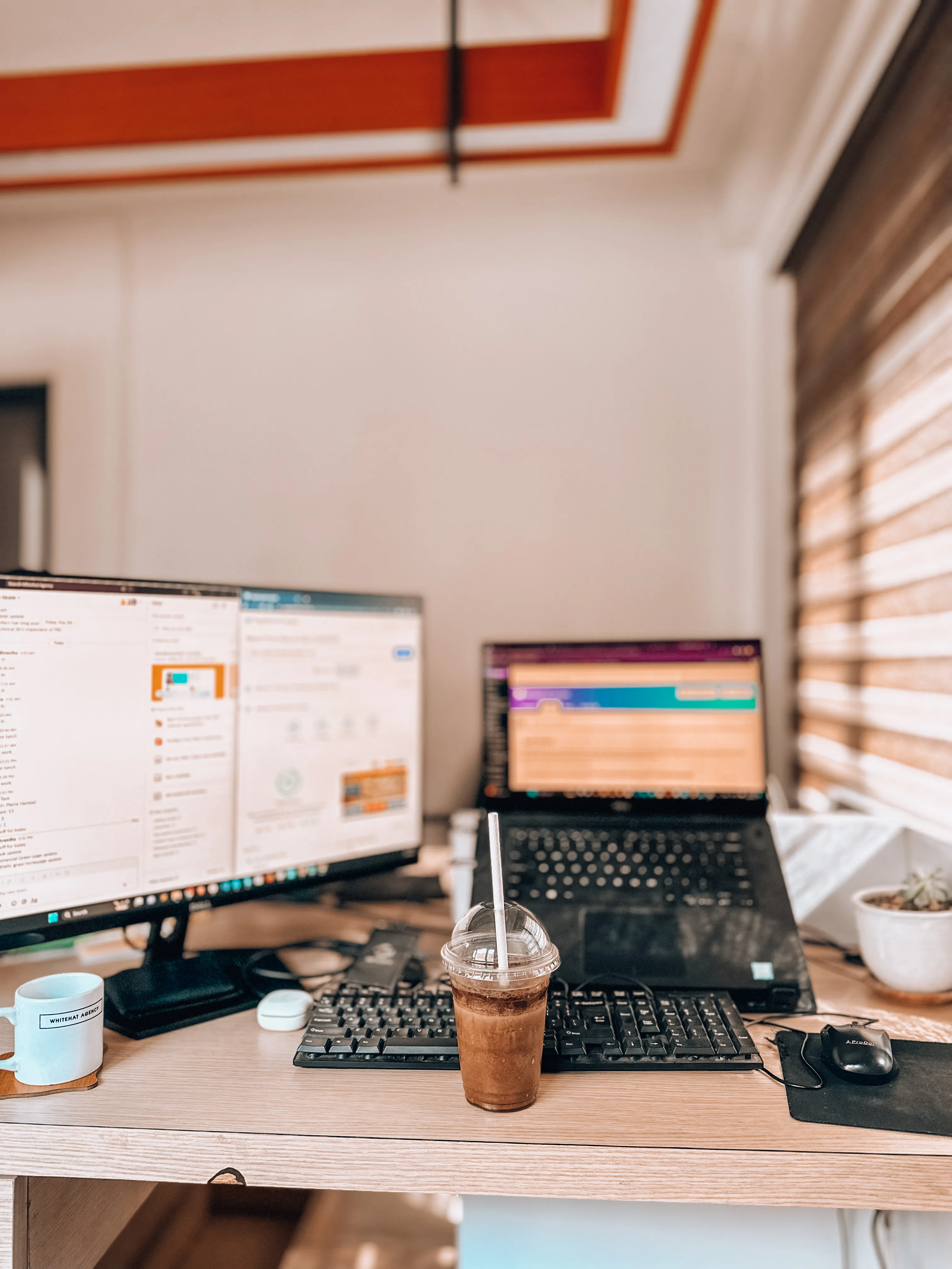 Laptop and desktop monitor on a wooden desk beside a keyboard and iced drink