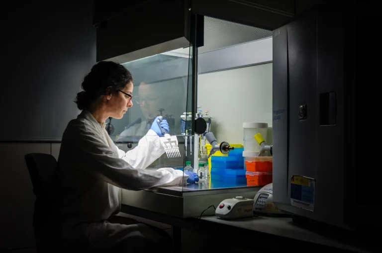 A researcher wearing glasses, a white laboratory coat and blue gloves uses a multichannel pipette at an illuminated laboratory workstation