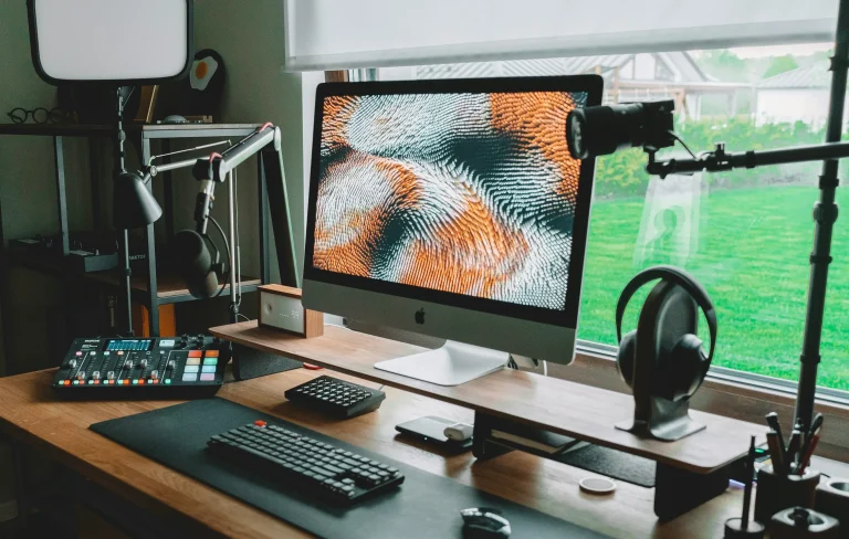 A desktop computer, keyboard, headphones and audio equipment sit on a wooden desk beside a window