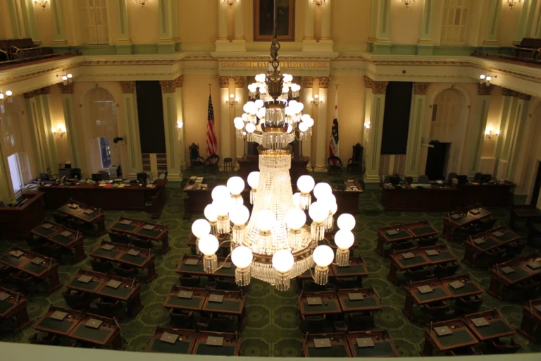 Chandeliers above rows of desks in the California State Assembly chamber, with the speaker’s dais and flags beyond