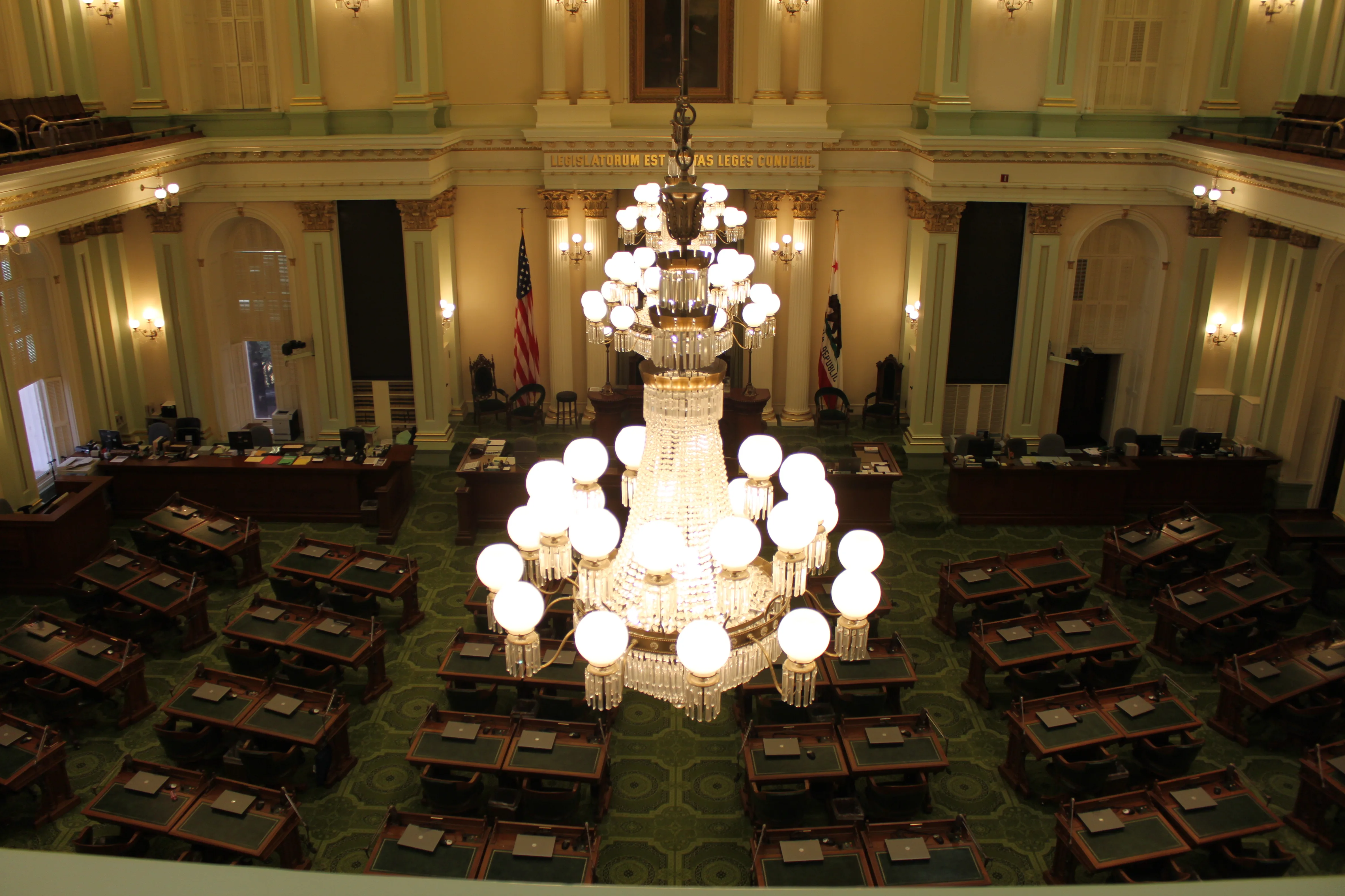 Chandeliers above rows of desks in the California State Assembly chamber, with the speaker’s dais and flags beyond