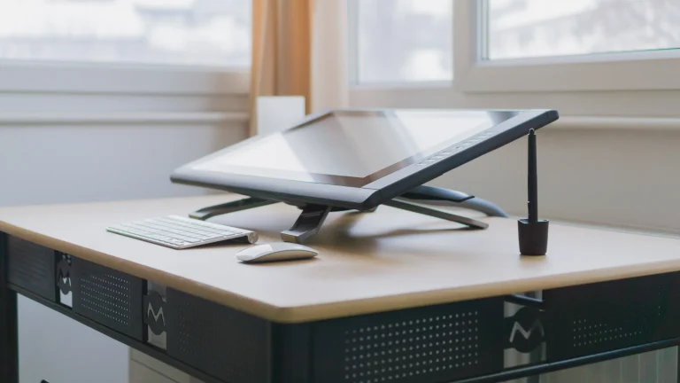 A Wacom drawing display on an angled stand beside a black stylus, white keyboard and mouse near a bright window