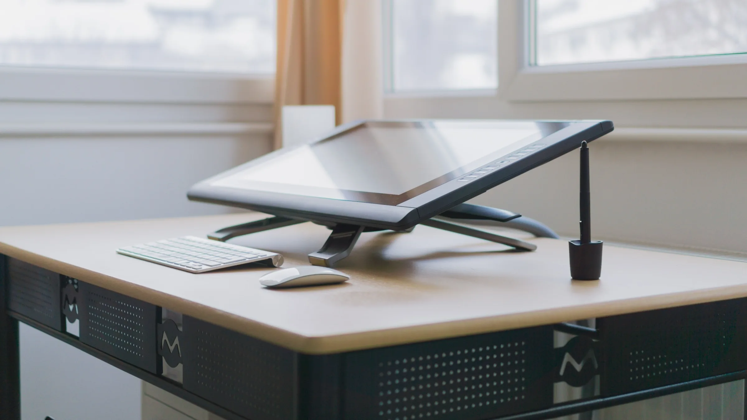 A Wacom drawing display on an angled stand beside a black stylus, white keyboard and mouse near a bright window