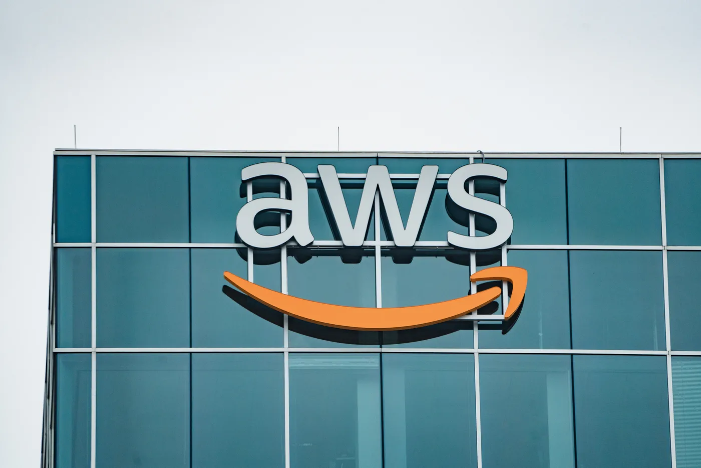 White AWS letters and an orange smile logo mounted on a glass office facade beneath a pale sky
