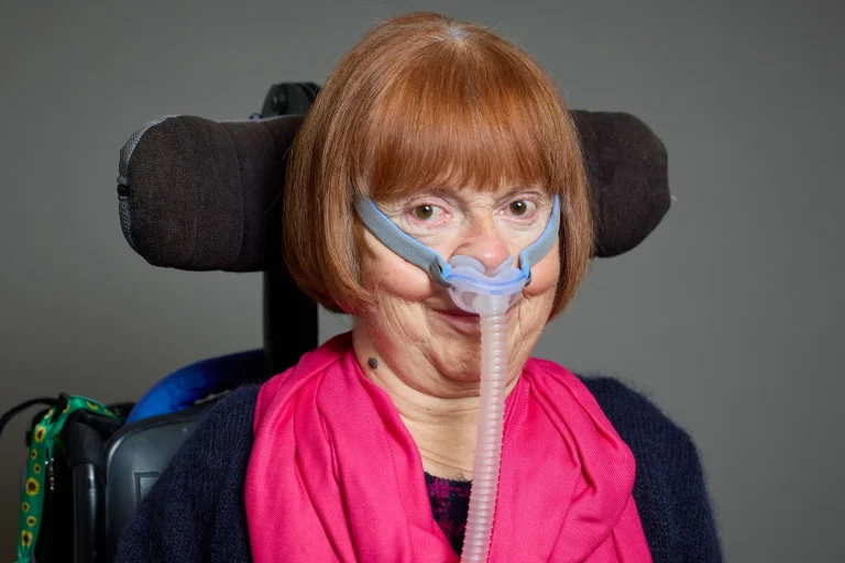Official portrait of Baroness Campbell of Surbiton wearing a pink scarf and seated in her wheelchair.