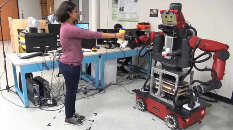 A person holds a cup toward the gripper of a red Baxter robot in a laboratory