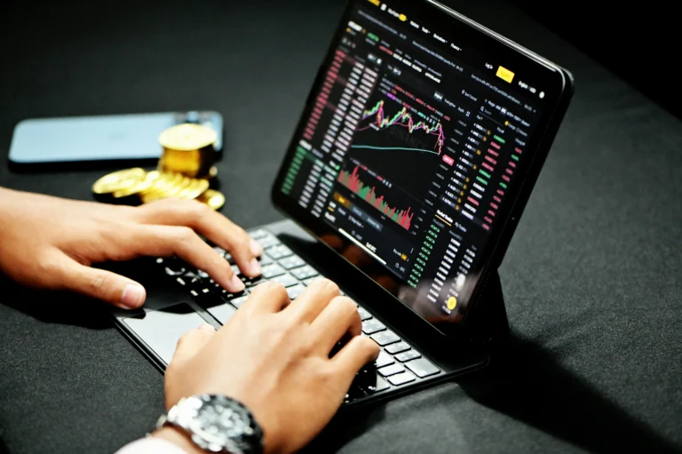 Hands on a keyboard beside a tablet displaying the Binance trading platform, with a phone and gold colored tokens nearby