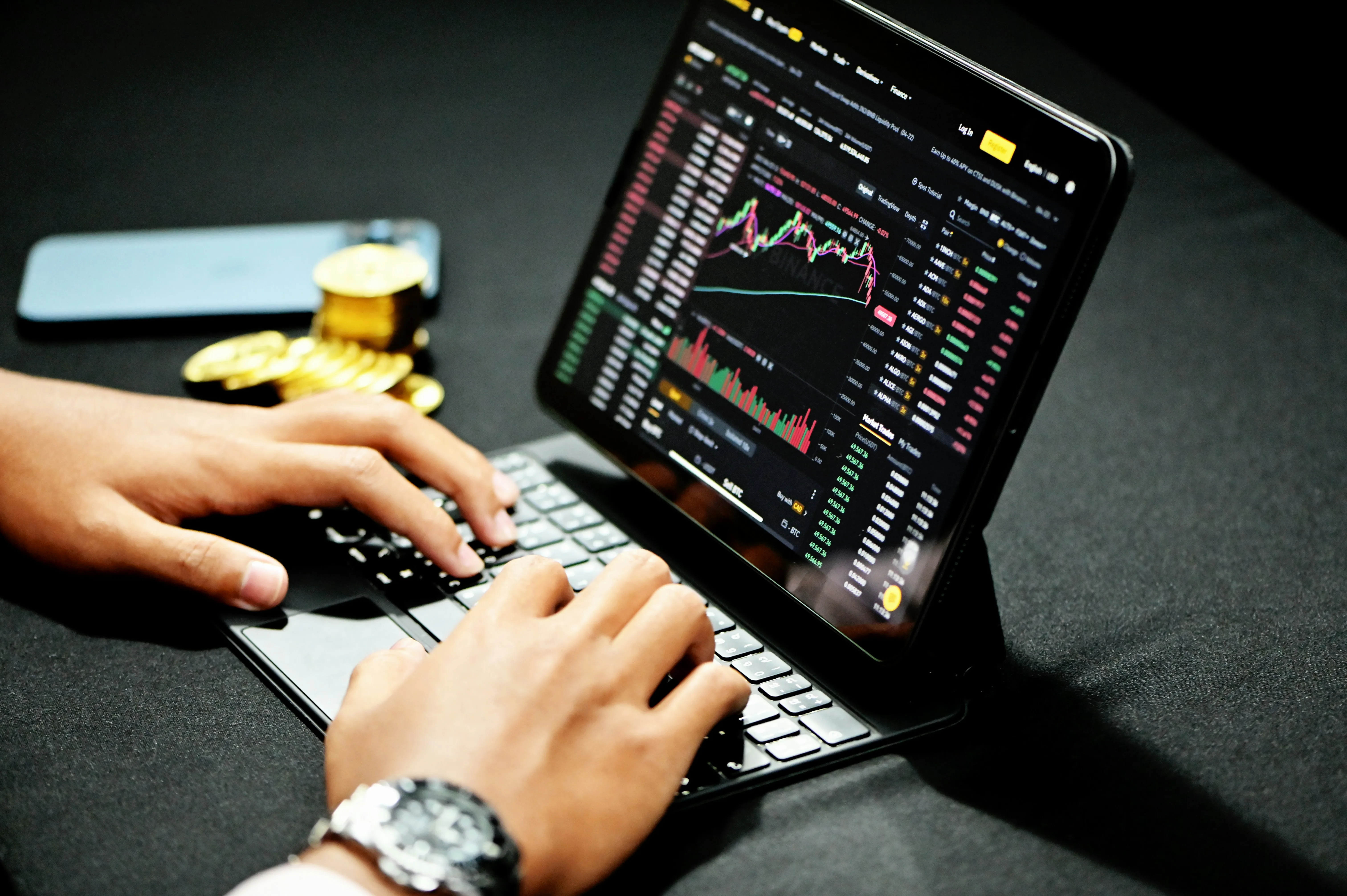 Hands on a keyboard beside a tablet displaying the Binance trading platform, with a phone and gold colored tokens nearby