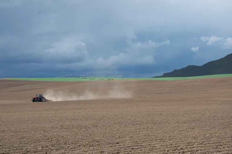 A tractor raises a trail of dust in an open field beneath dark clouds