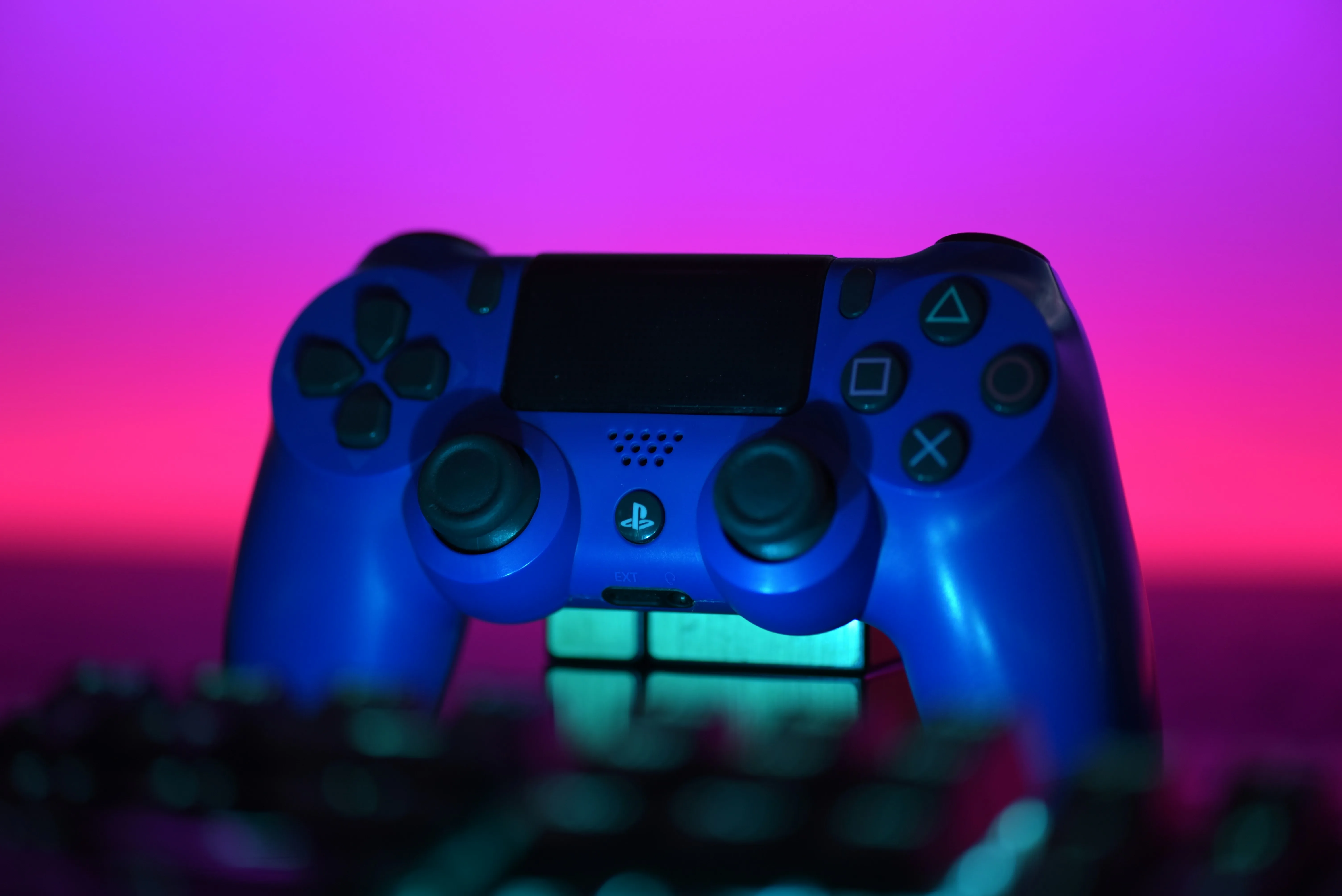 Blue PlayStation 4 controller behind a blurred keyboard under pink and purple lighting