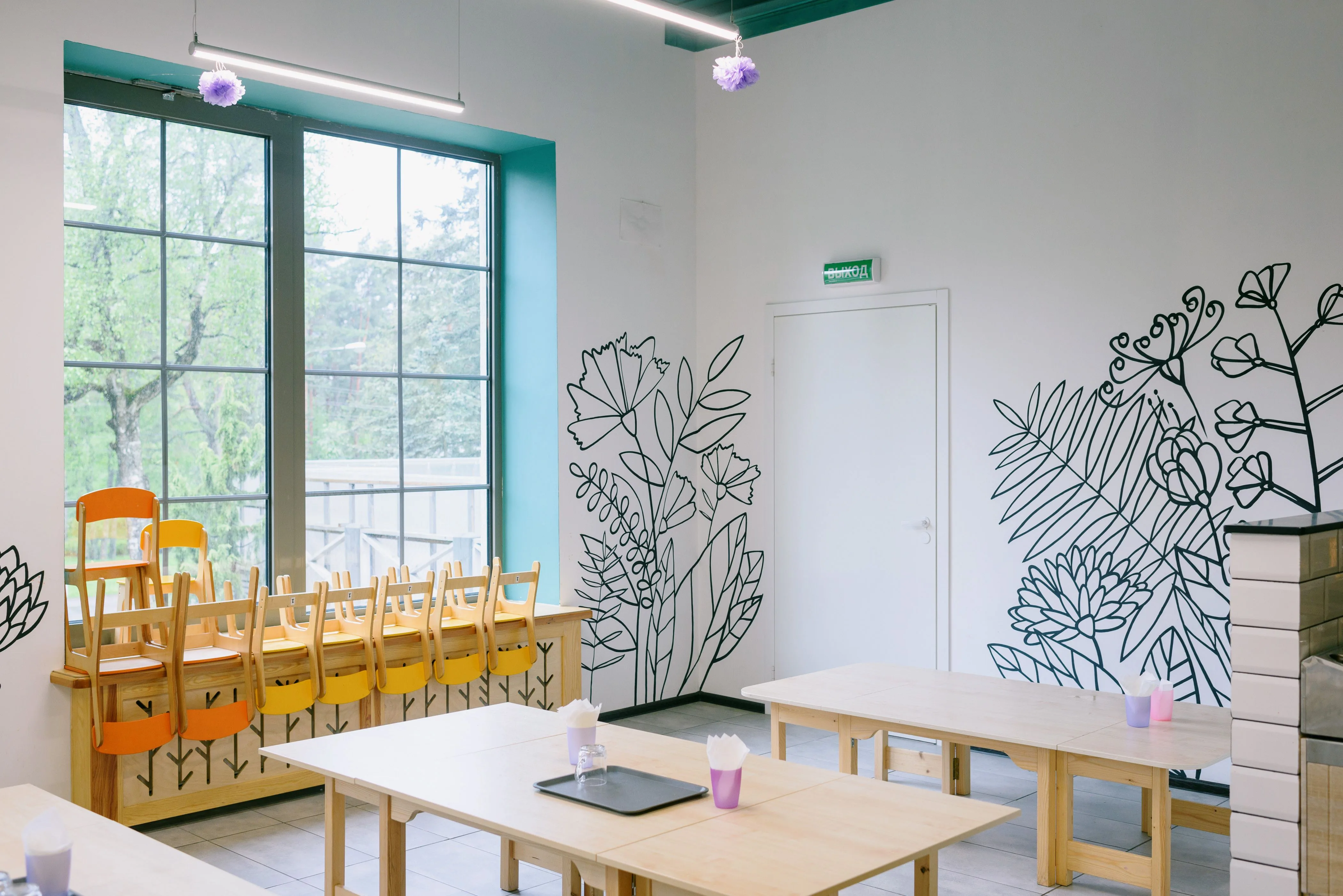 An empty classroom with wooden tables and chairs beside large windows and floral wall drawings