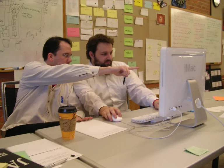 Two programmers sharing an iMac workstation with one pointing at the display