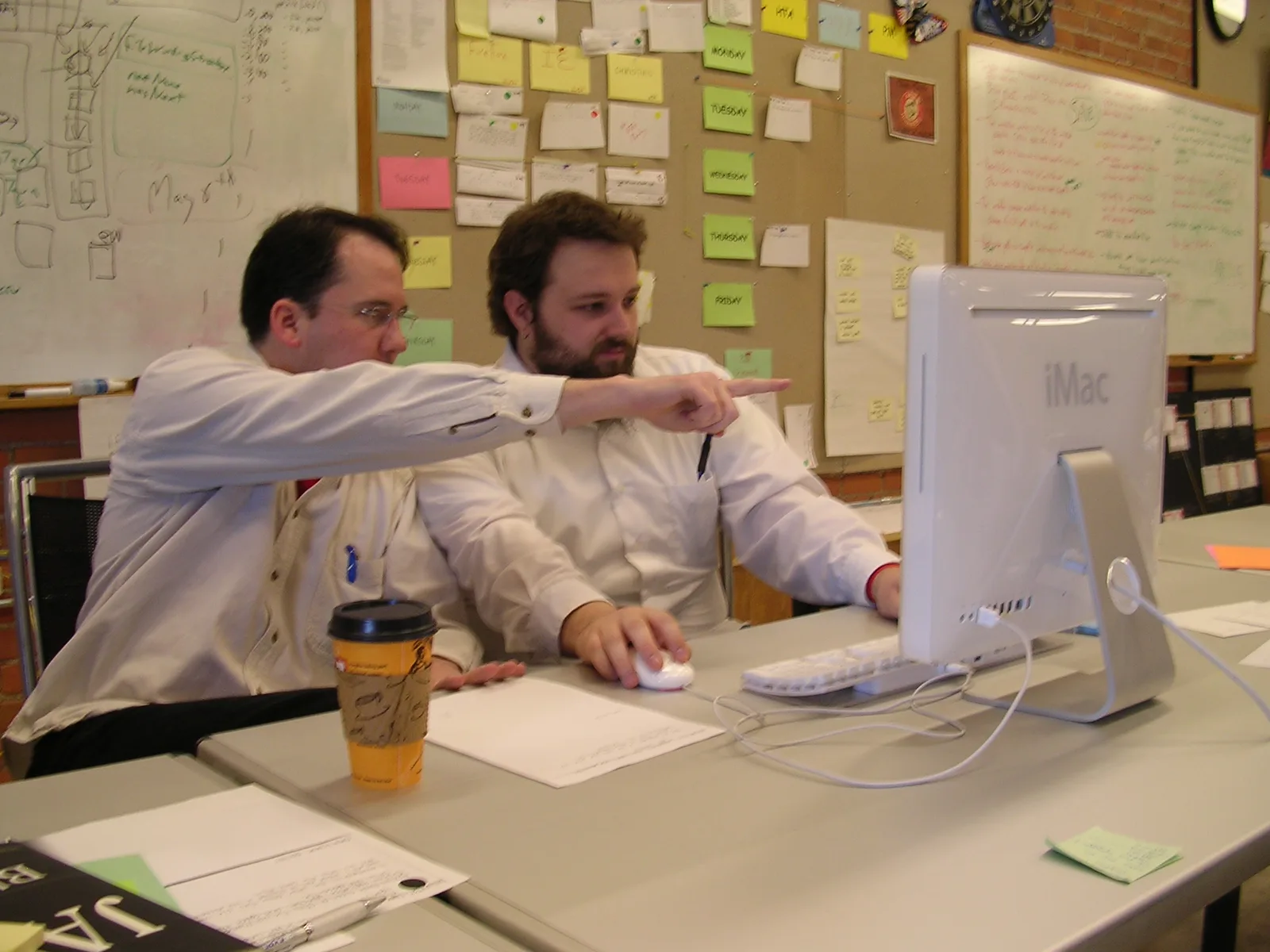 Two programmers sharing an iMac workstation with one pointing at the display
