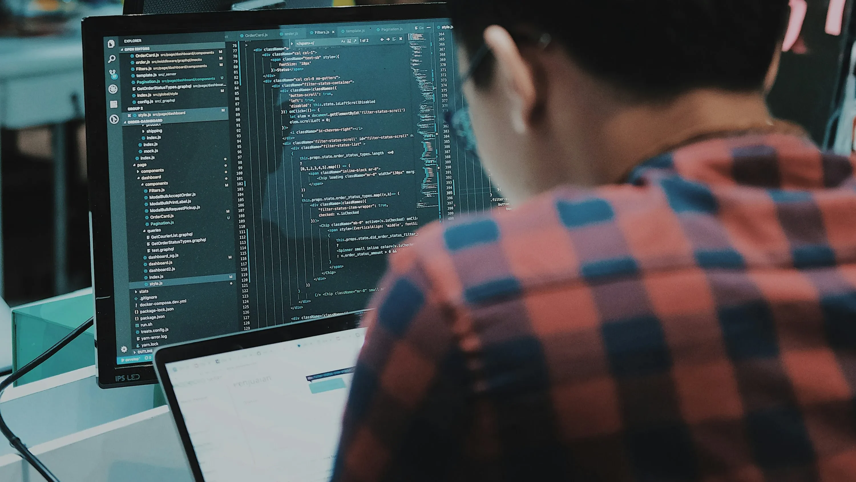 A developer in a plaid shirt faces a monitor displaying source code in a shared workspace