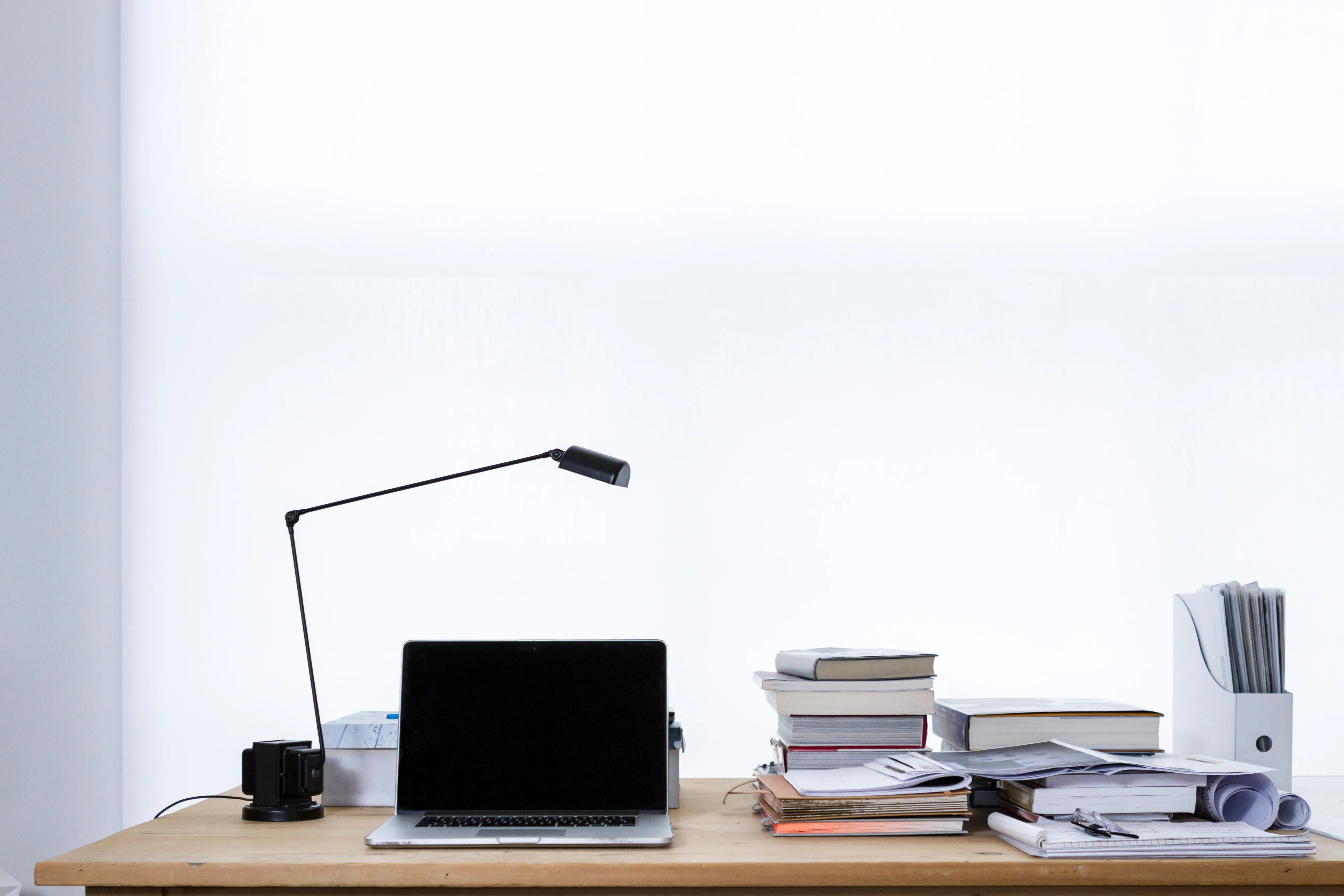 A laptop with a dark screen sits beside books and papers beneath a black desk lamp