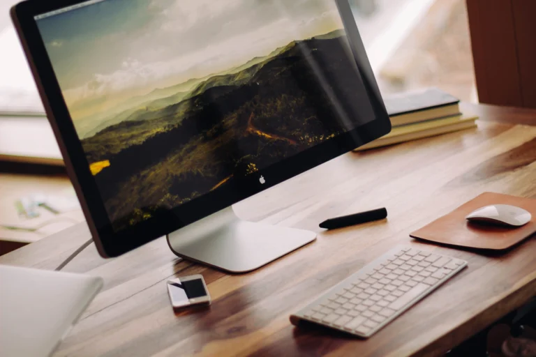 A desktop monitor showing a mountain landscape beside a keyboard and mouse on a wooden desk