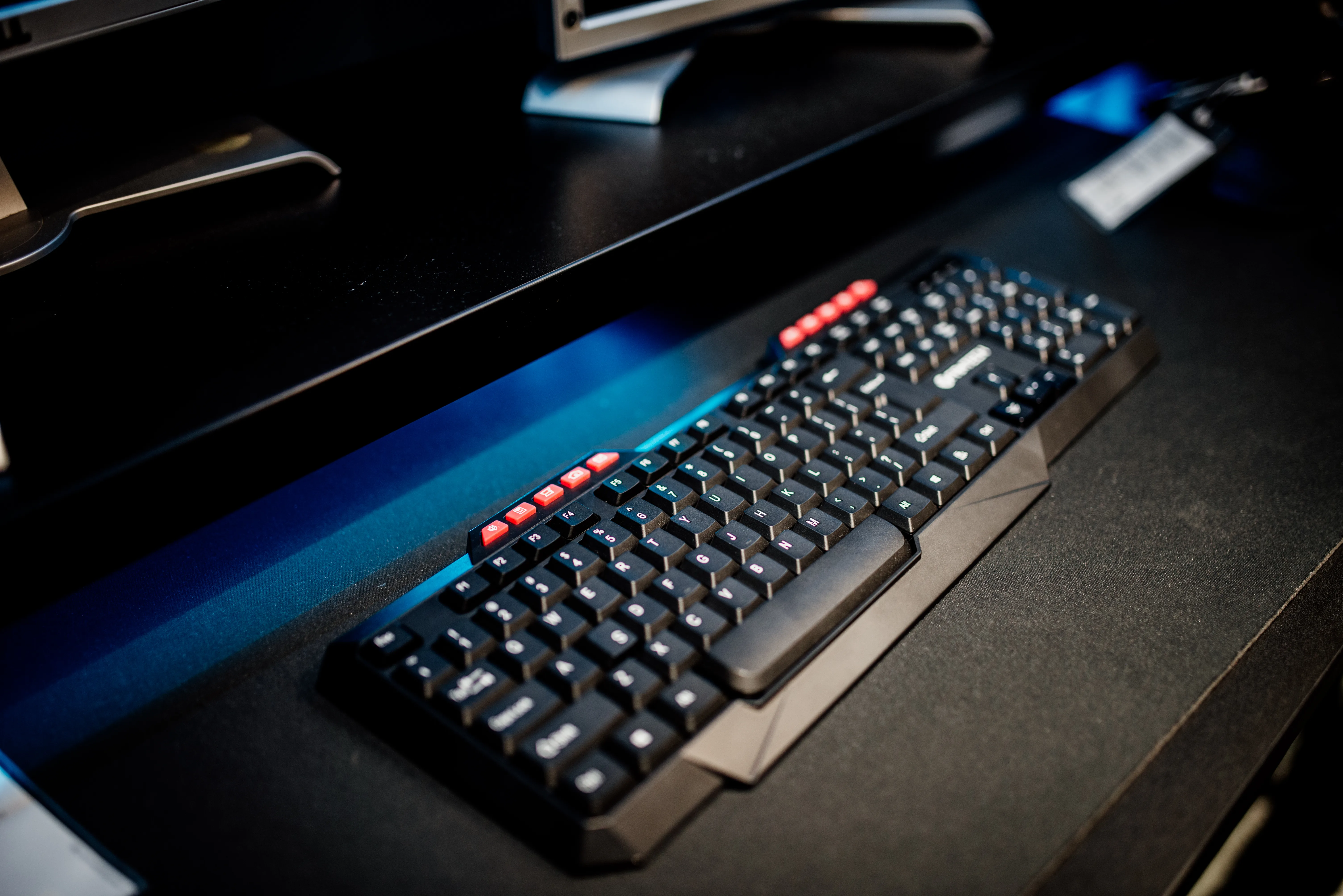 Black keyboard with red keys on a desk beneath desktop monitors