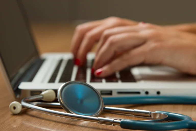 Stethoscope on a wooden desk beside hands typing on a laptop