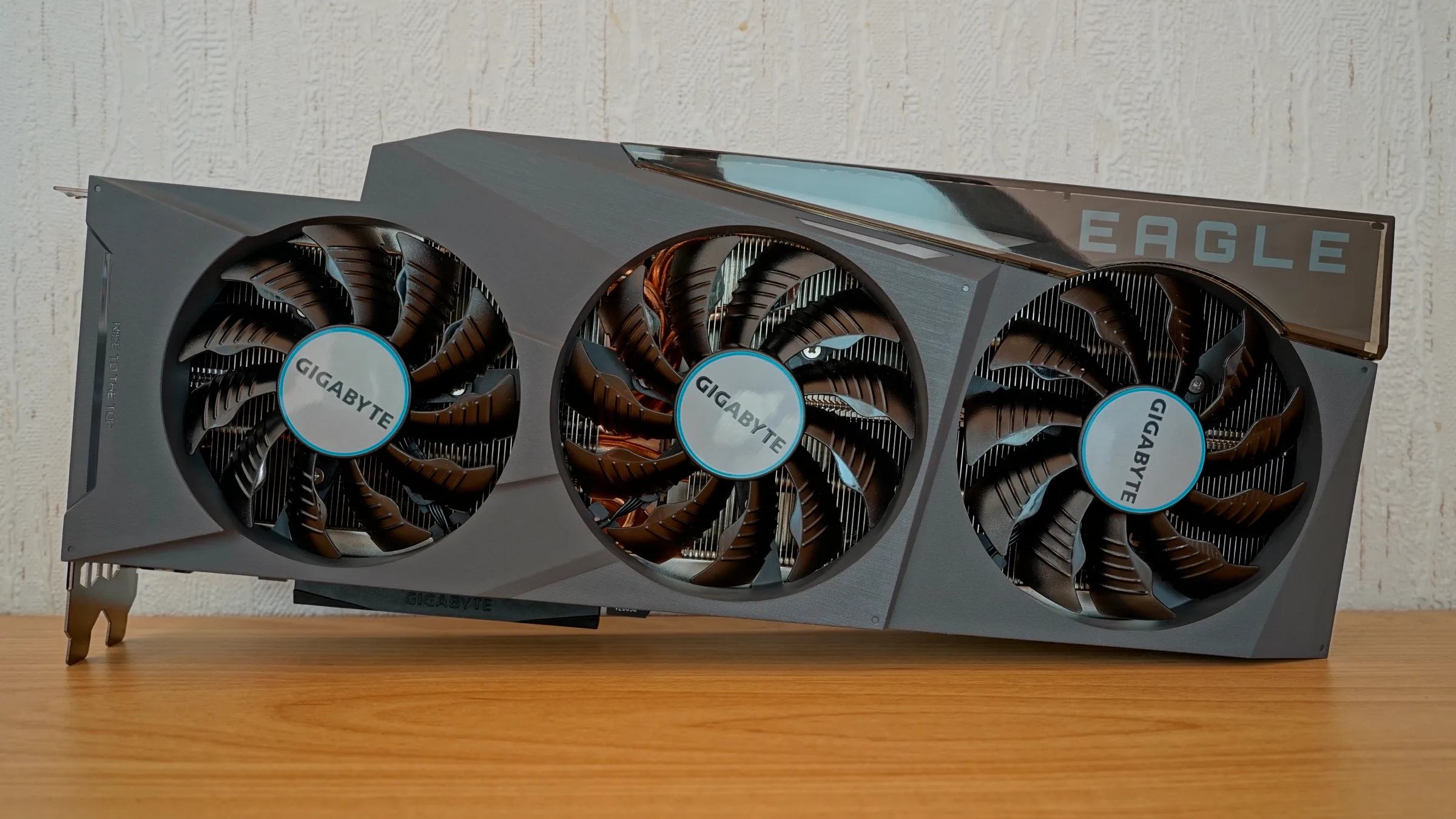 Three black cooling fans on a gray Gigabyte GeForce RTX 3090 Eagle graphics card resting on a wooden surface