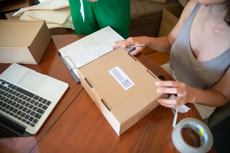 Hands hold a cardboard parcel beside a paper ledger, pencil and laptop on a wooden packing table