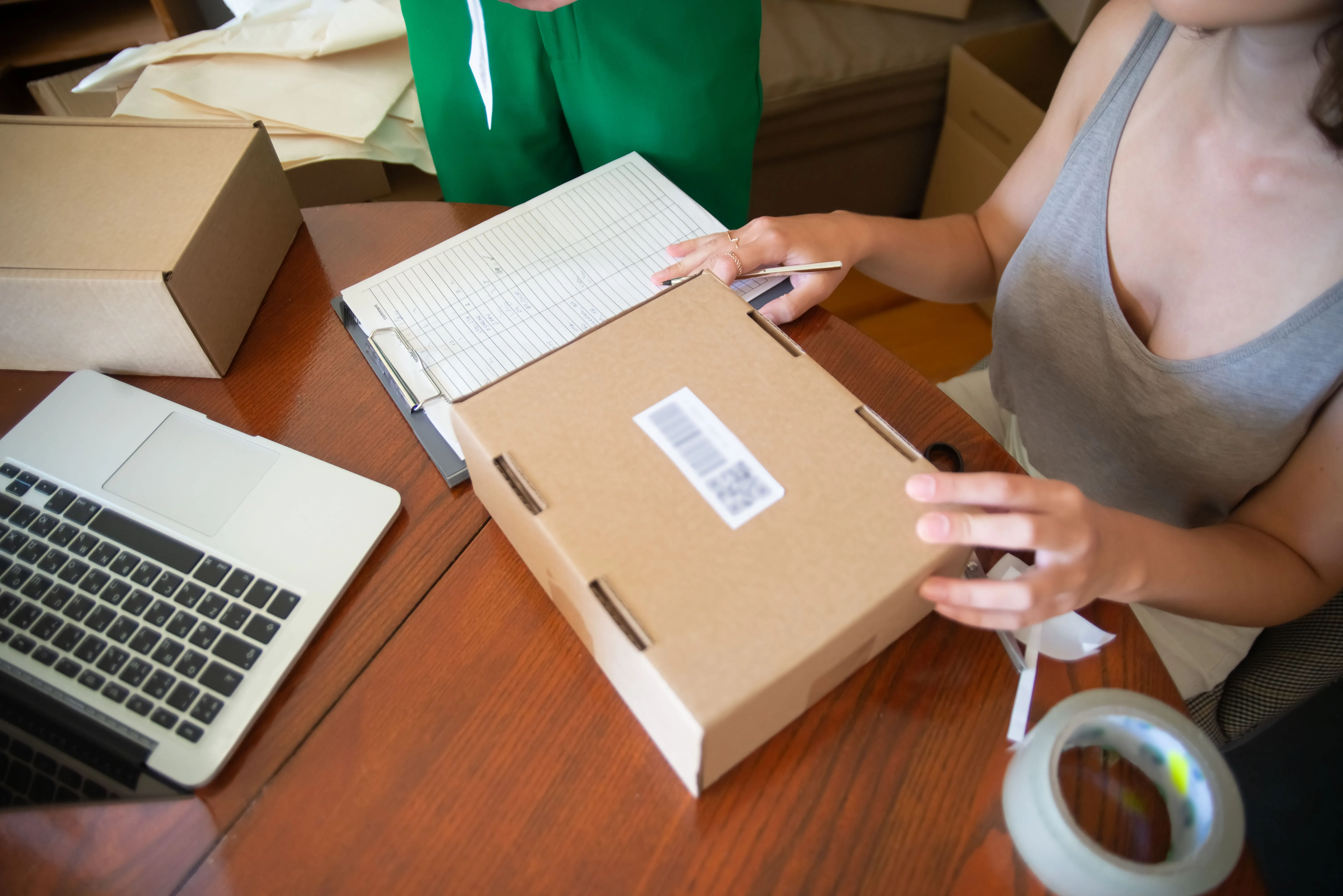 Hands hold a cardboard parcel beside a paper ledger, pencil and laptop on a wooden packing table