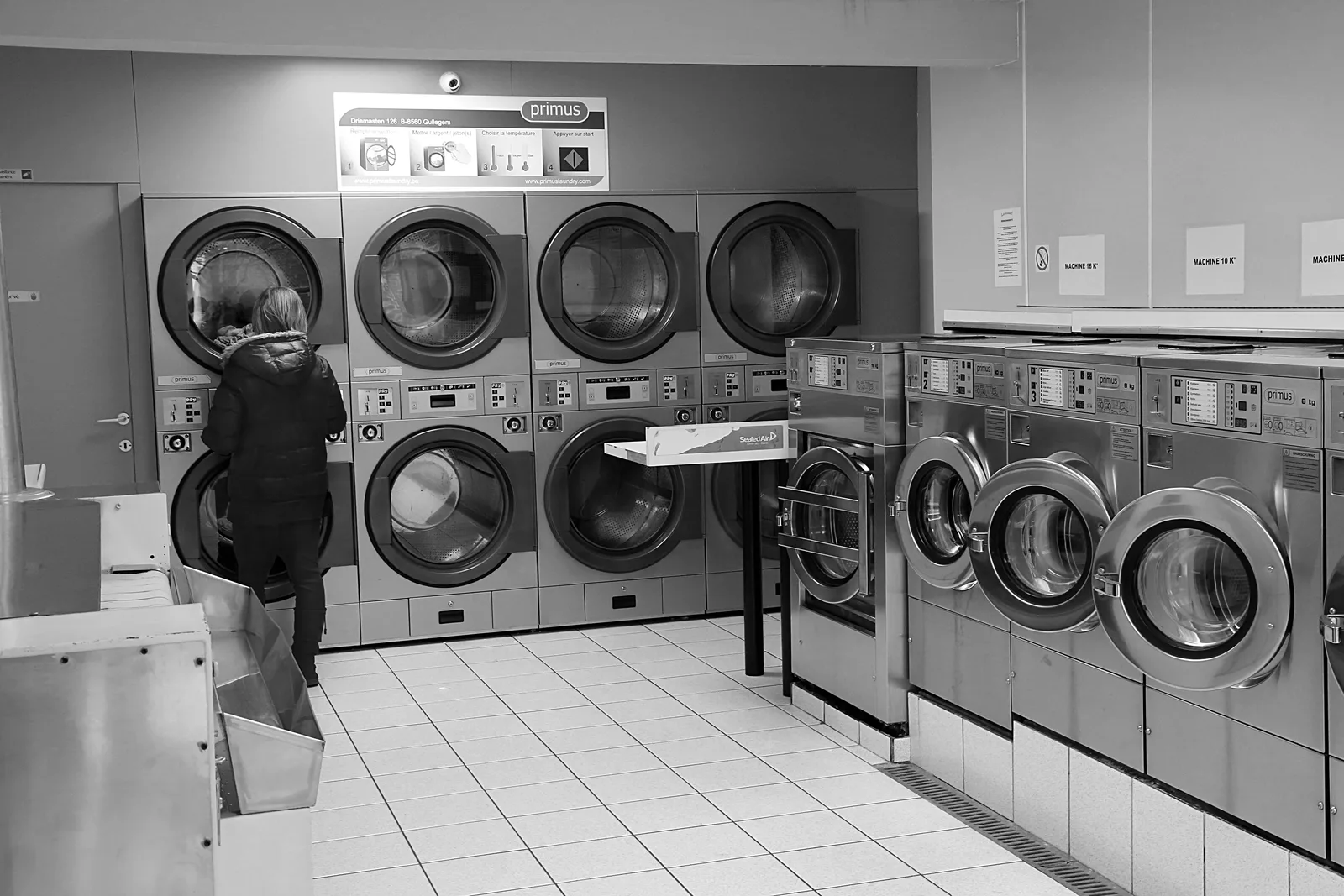 Rows of washing machines and dryers with a customer facing a dryer in a Belgian laundromat