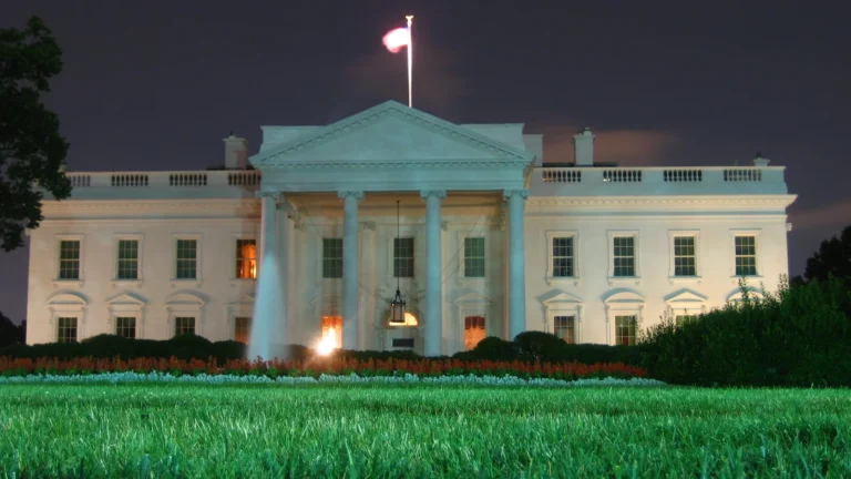 Illuminated north facade of the White House behind a lawn beneath a dark sky