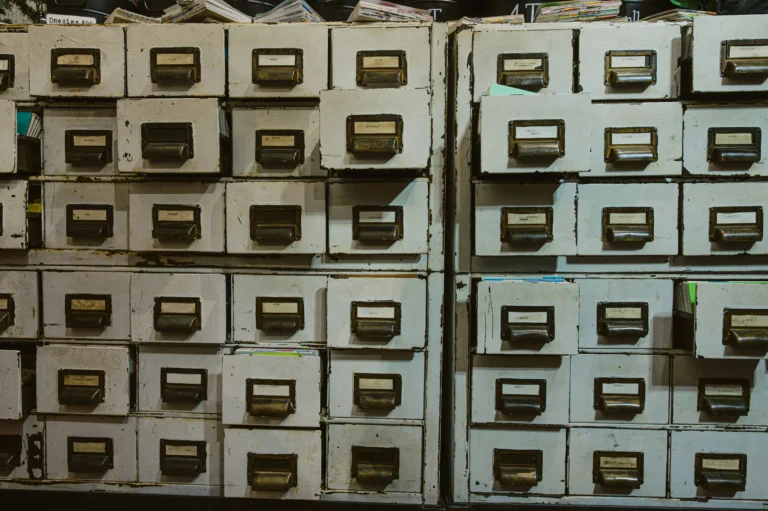 Rows of worn white filing drawers with metal handles and labels, with papers in some open drawers