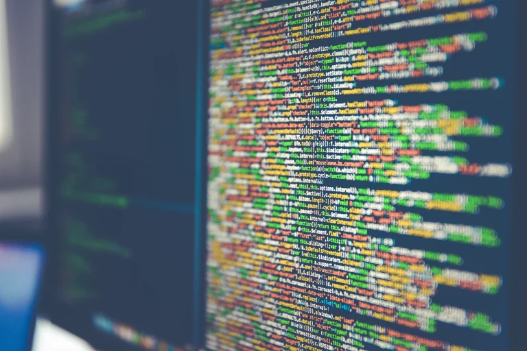 Colorful source code on an angled dark computer monitor, with a softly blurred second screen to the left.