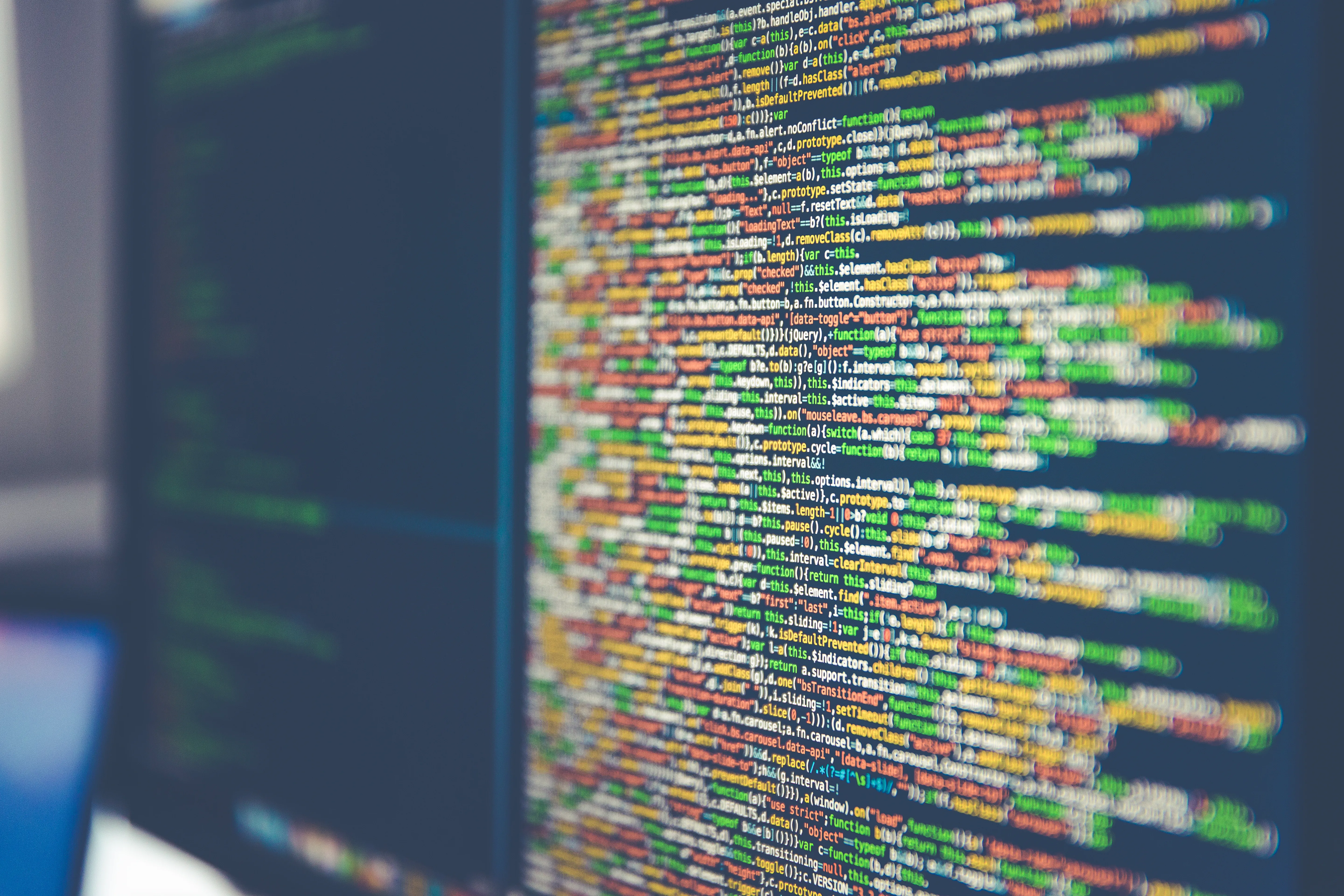 Colorful source code on an angled dark computer monitor, with a softly blurred second screen to the left.