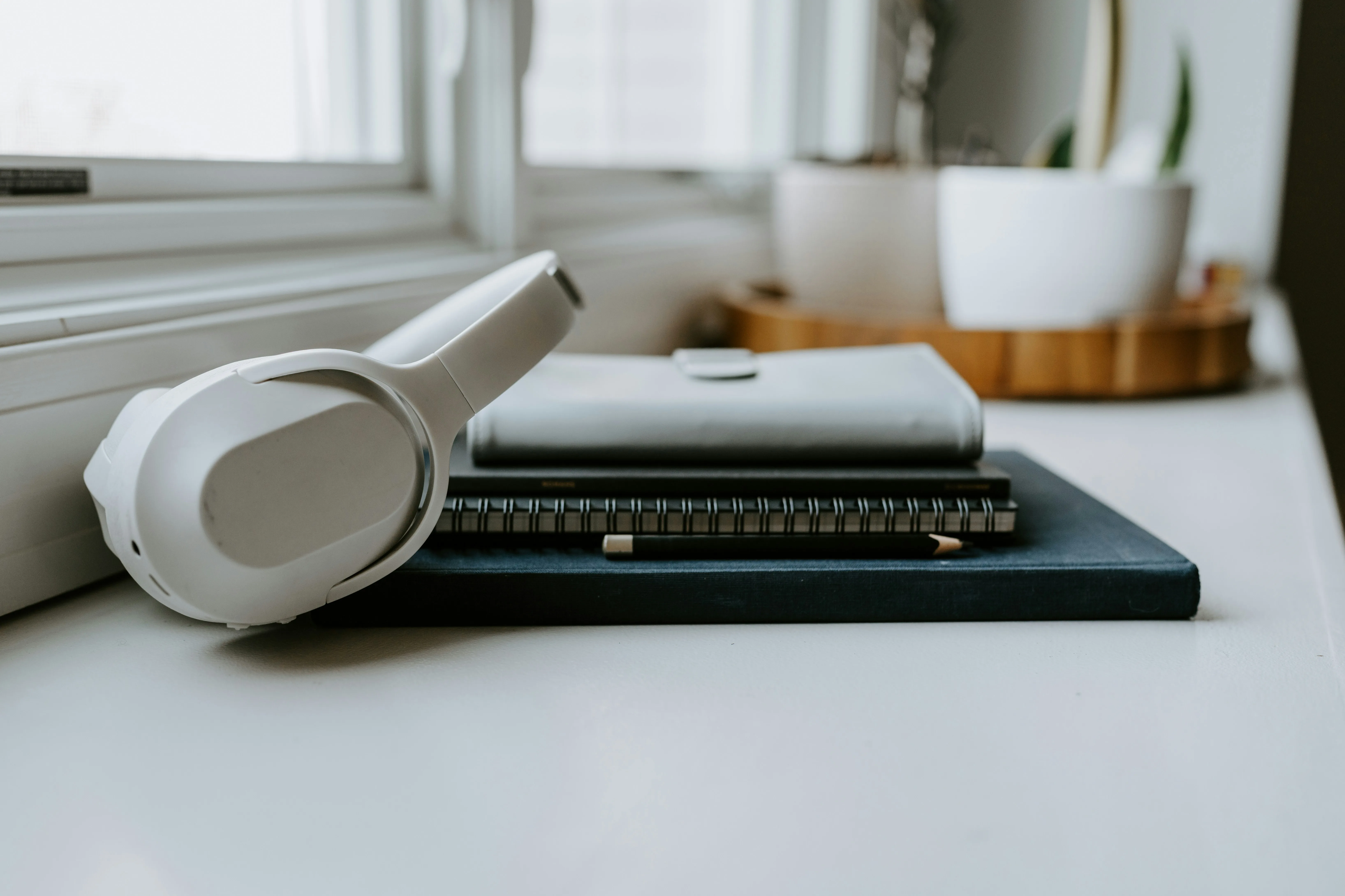 White headphones beside stacked books and notebooks with a pencil on a windowsill