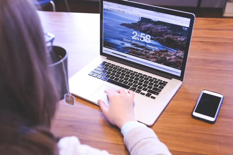 A person uses the trackpad of a silver laptop displaying a Chrome new tab page on a wooden table beside a mug and phone