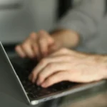 Hands type on a laptop keyboard with a wristwatch visible and a blurred grey background