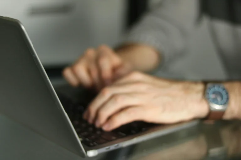 Hands type on a laptop keyboard with a wristwatch visible and a blurred grey background
