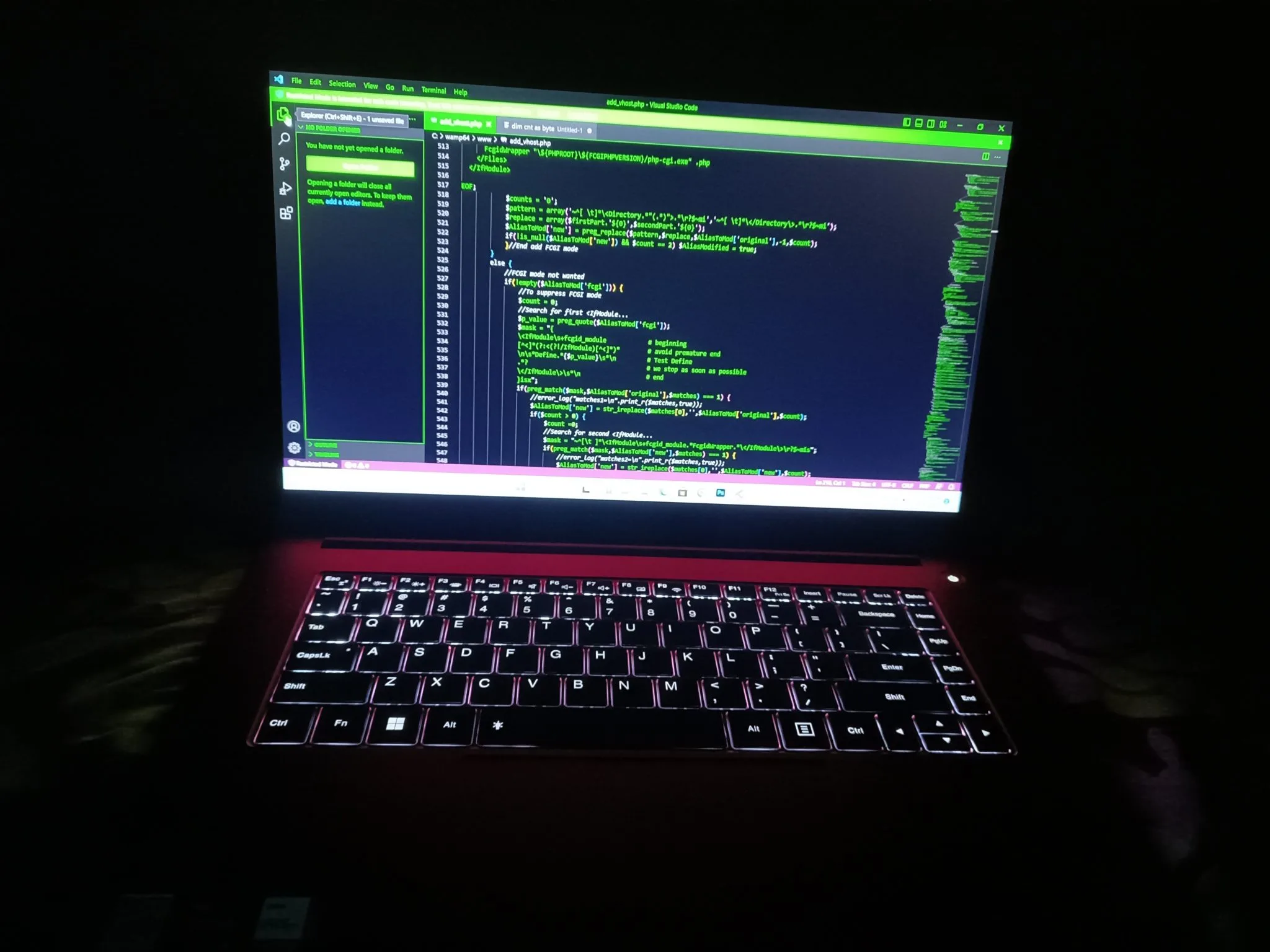A laptop displays code in green above an illuminated keyboard in a dark room.