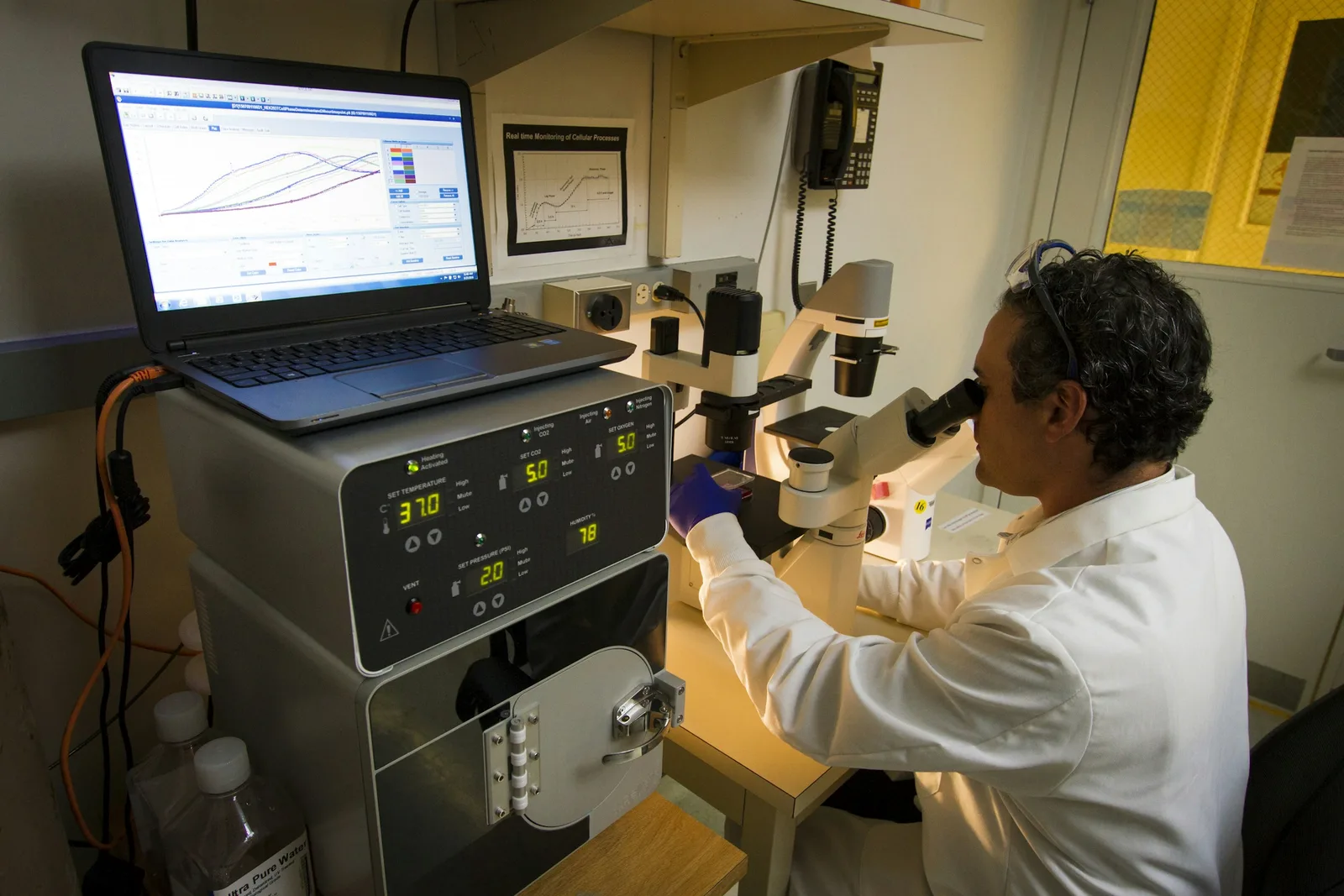 Technician using a microscope beside a computer at the Cancer Genomics Research Laboratory