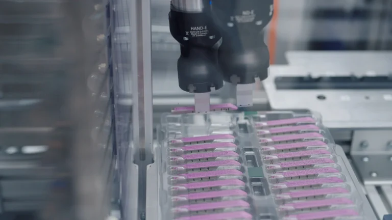 Robot gripper above rows of purple components in a manufacturing enclosure.