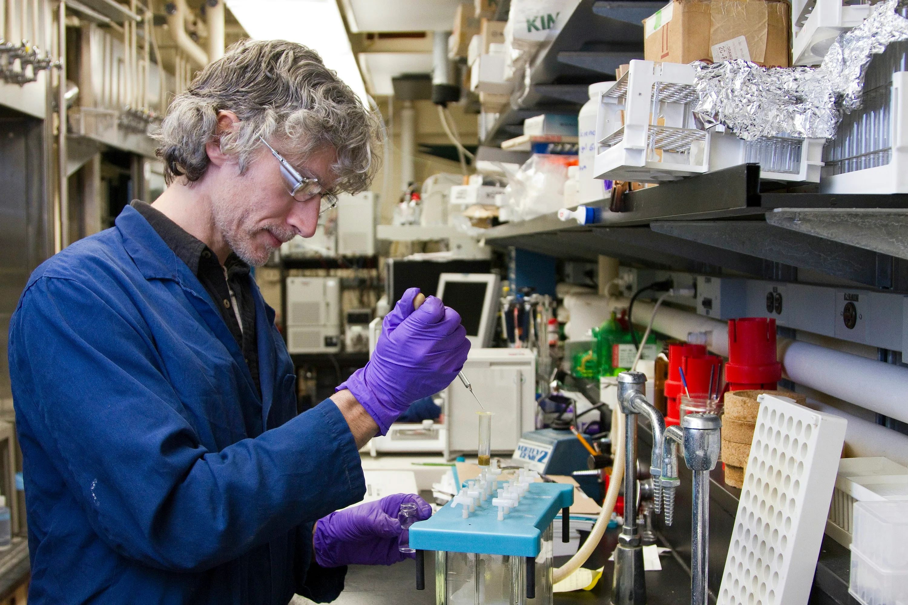 Researcher wearing a blue laboratory coat and purple gloves pipetting into a tube at a laboratory bench
