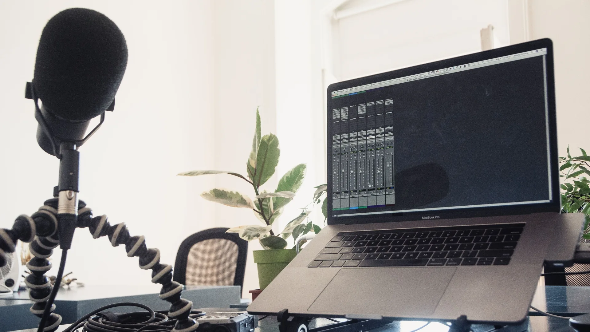 A black microphone on a flexible tripod beside a laptop displaying audio mixing software on a bright desk