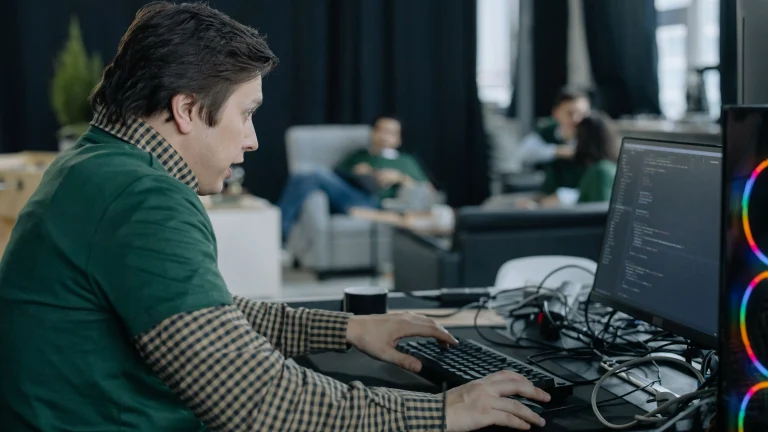 A man in a green shirt works at a keyboard and mouse beside a monitor displaying softly focused source code