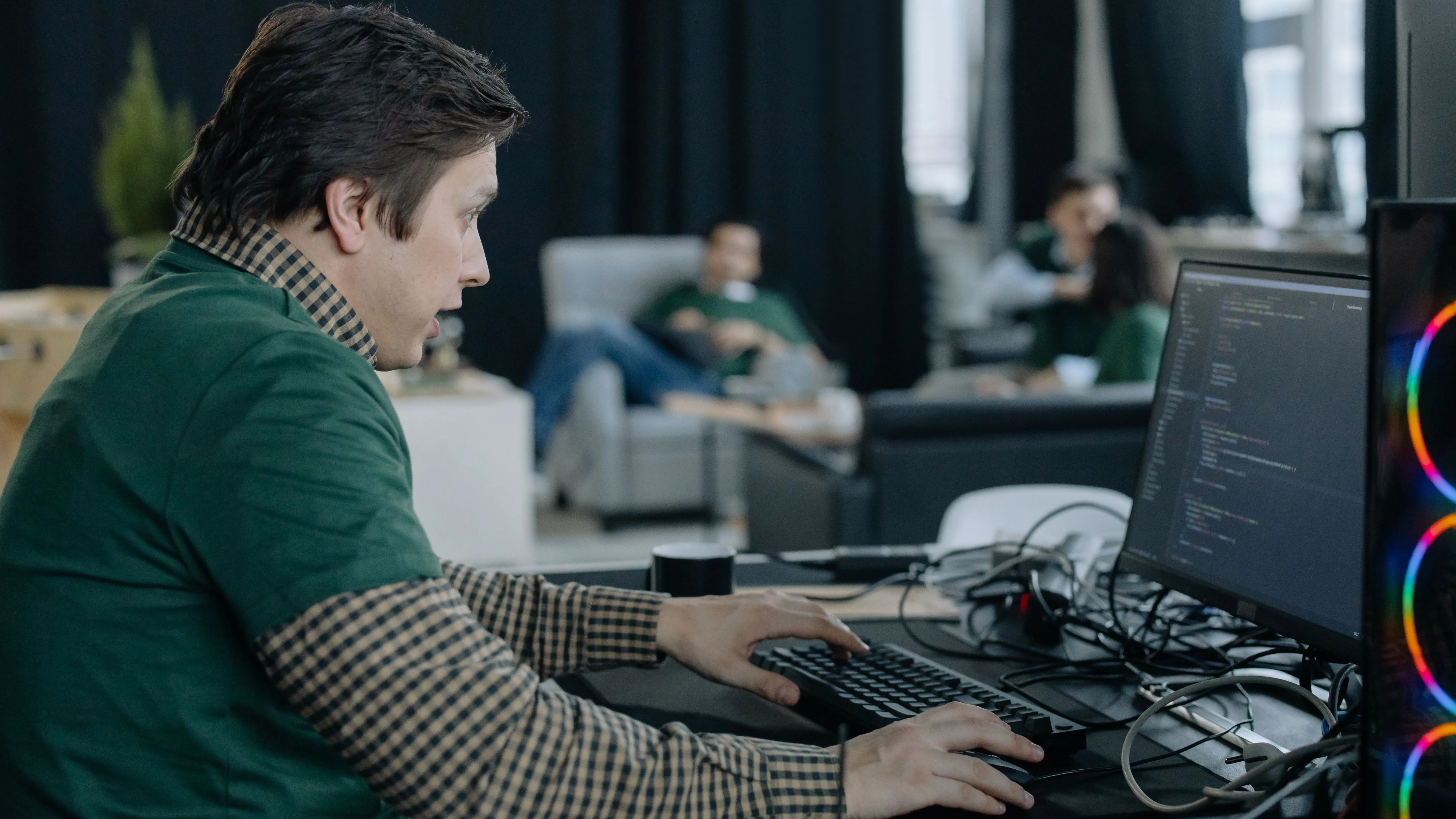 A man in a green shirt works at a keyboard and mouse beside a monitor displaying softly focused source code
