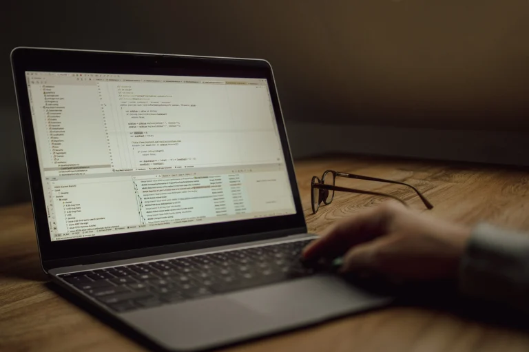 Hands at a laptop displaying programming code with eyeglasses on the wooden desk