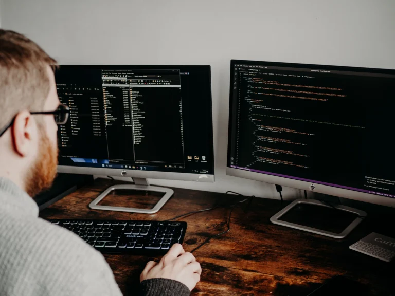 A developer works at two monitors displaying code on a wooden desk