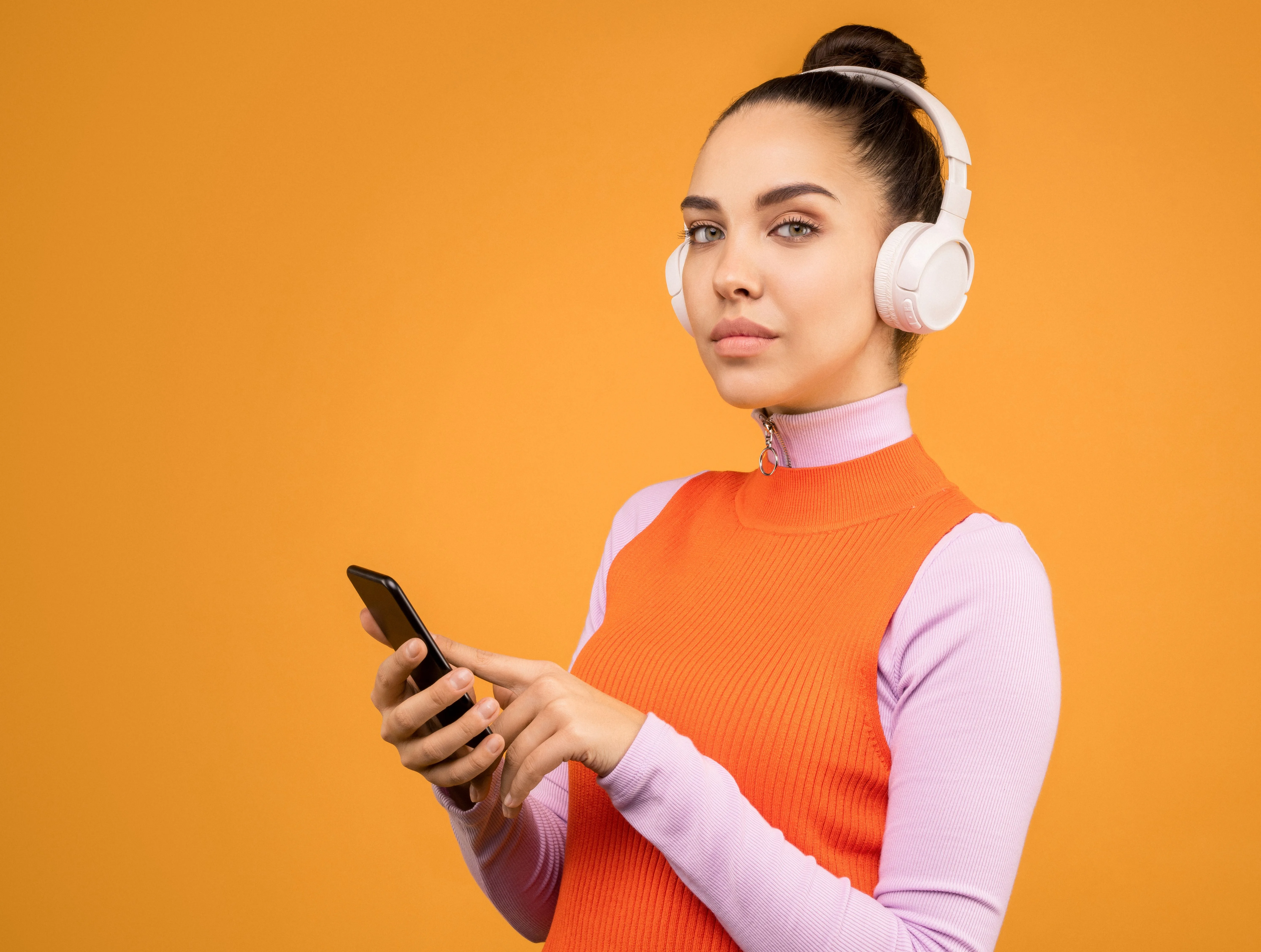 A woman wearing white headphones holds a smartphone against an orange background