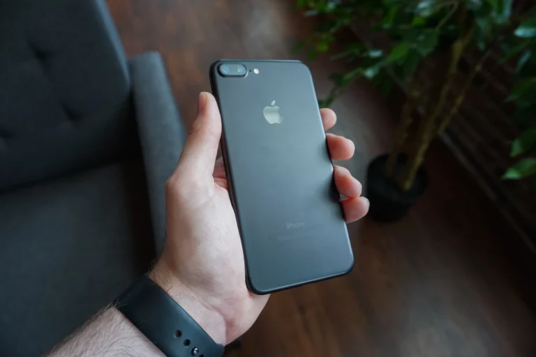 A hand holds the back of a black iPhone beside a chair and a leafy plant