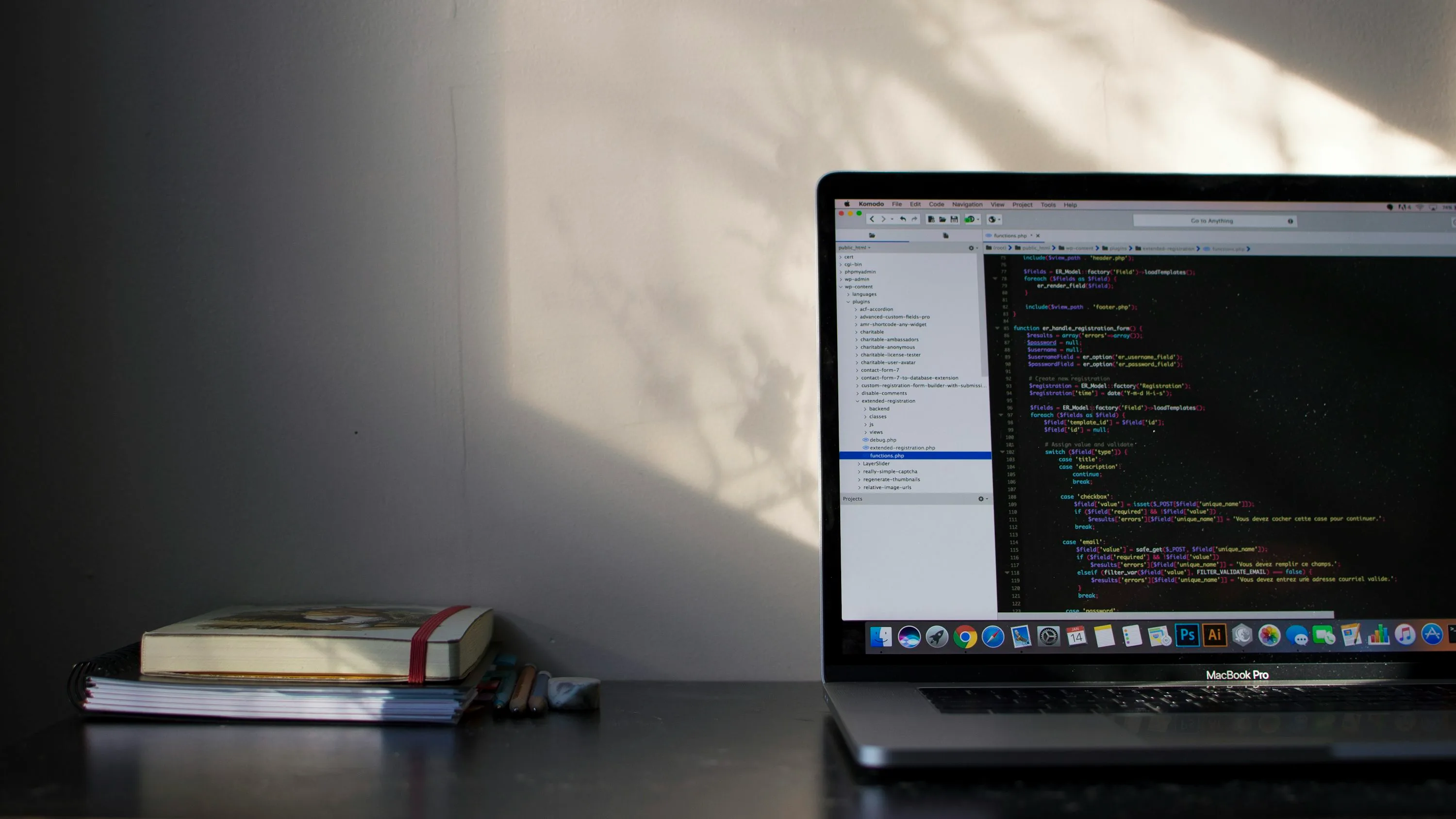 A laptop displaying source code sits beside a stack of notebooks in warm window light