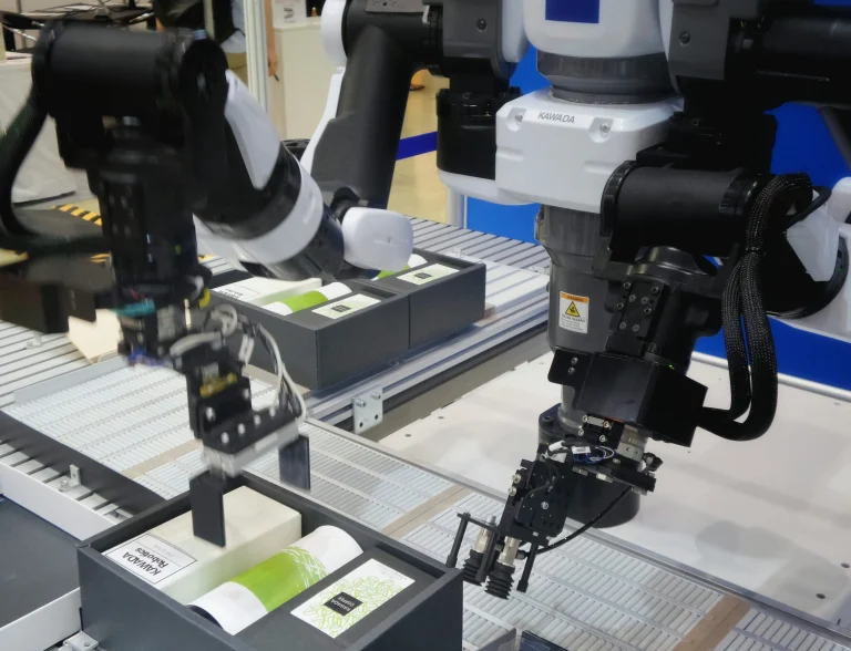 Two black and white robotic arms with grippers positioned above divided trays holding small boxes and containers