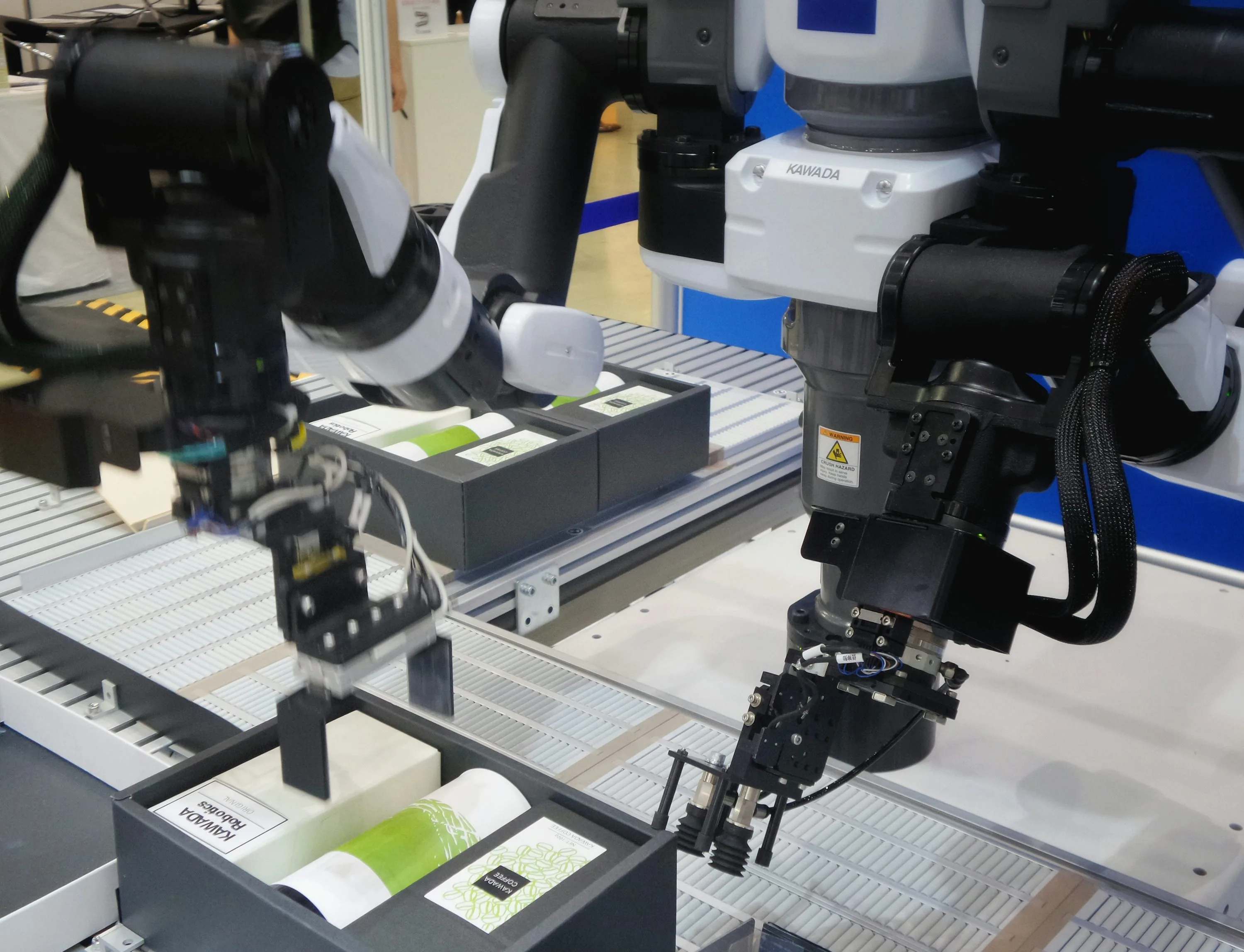 Two black and white robotic arms with grippers positioned above divided trays holding small boxes and containers