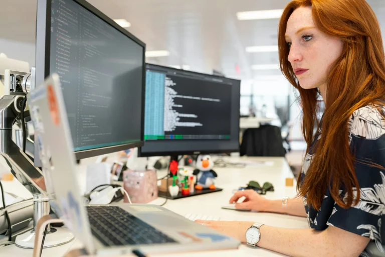 A person works at a desk beside two monitors displaying source code, with a laptop in the foreground
