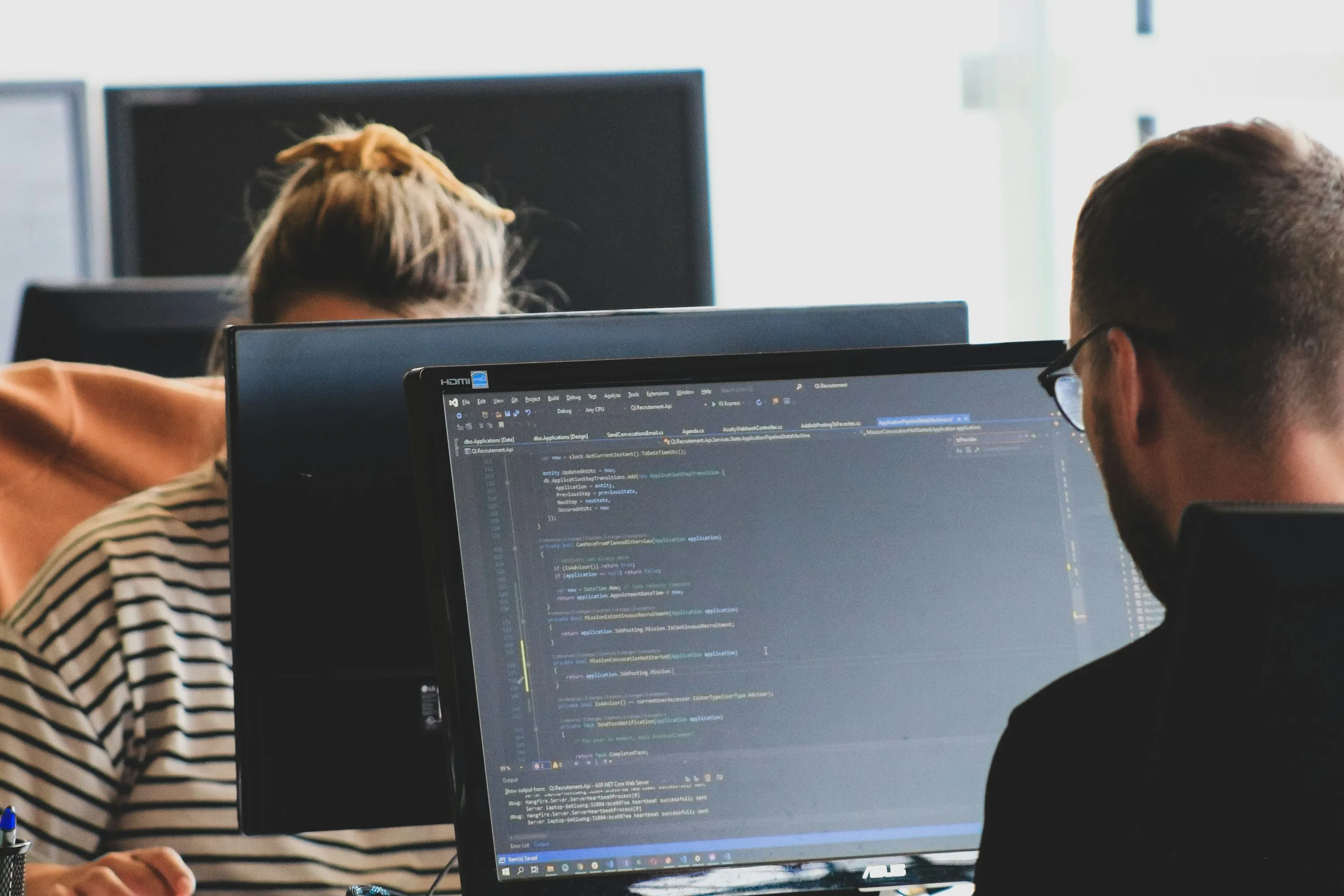 A person with glasses faces a monitor displaying source code and a console panel with another person behind a second screen